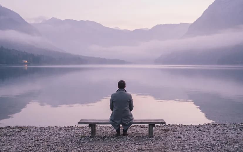 Eine Person sitzt von hinten gesehen auf einer Bank am See und schaut ins Abendlicht. (KI-Bild)