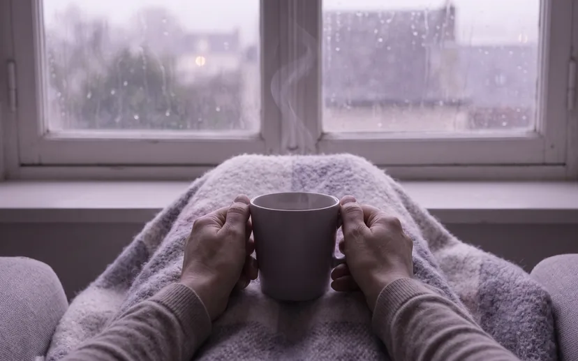 Zwei Hände halten eine warme Tasse, hinter dem Fenster laufen Regentropfen herunter. (KI-Bild)