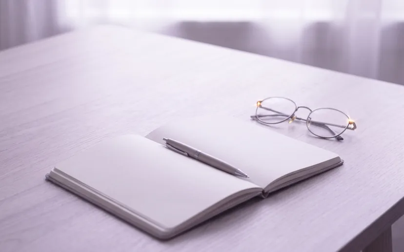 Ein offenes Notizbuch mit leerer Seite, darauf ein Stift, daneben eine Brille im Morgenlicht.