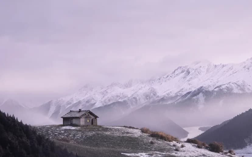 Ein einzelnes kleines Haus vor einer grossen, verschneiten Bergkette in der Dämmerung.