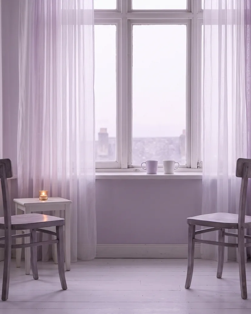 Zwei leere Stühle stehen am Fenster einander zugewandt, weiches Licht fällt herein.